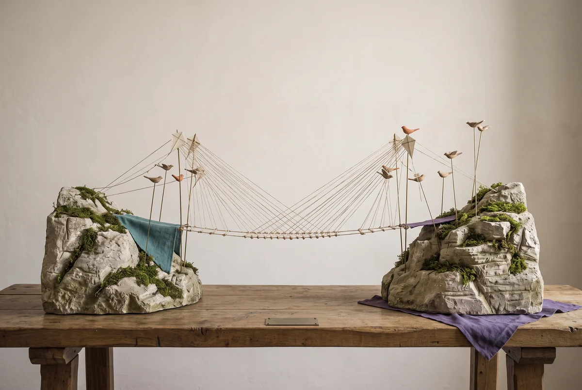 Drachen und Vögel weben eine Brücke aus Fäden zwischen zwei Hügeln.